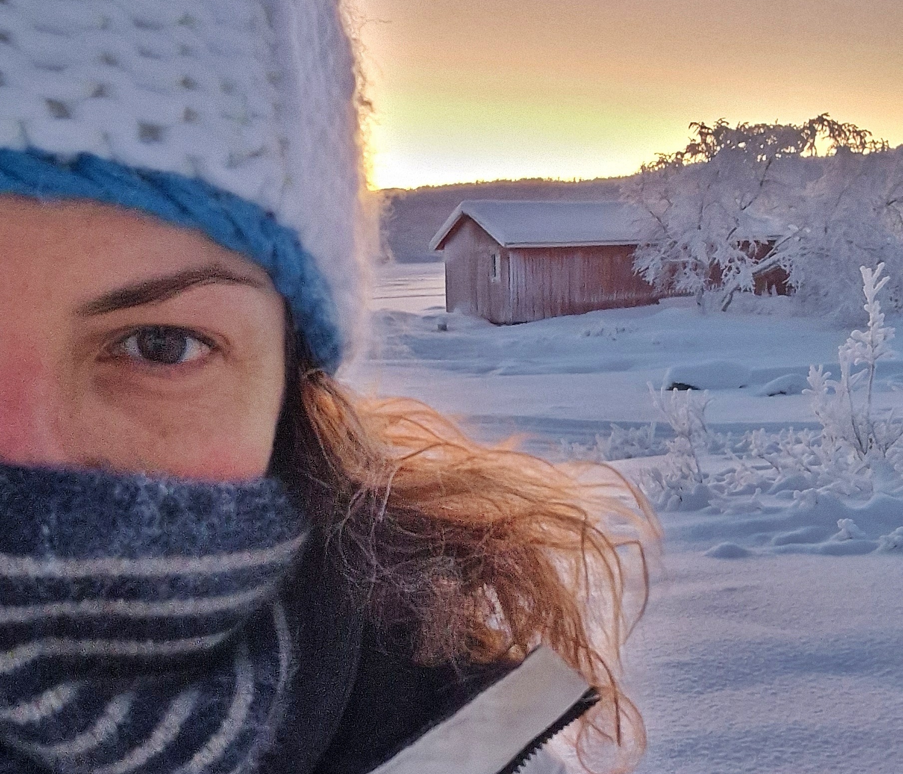 Persona con una casetta rossa di legno completamente innevata sullo sfondo, al tramonto